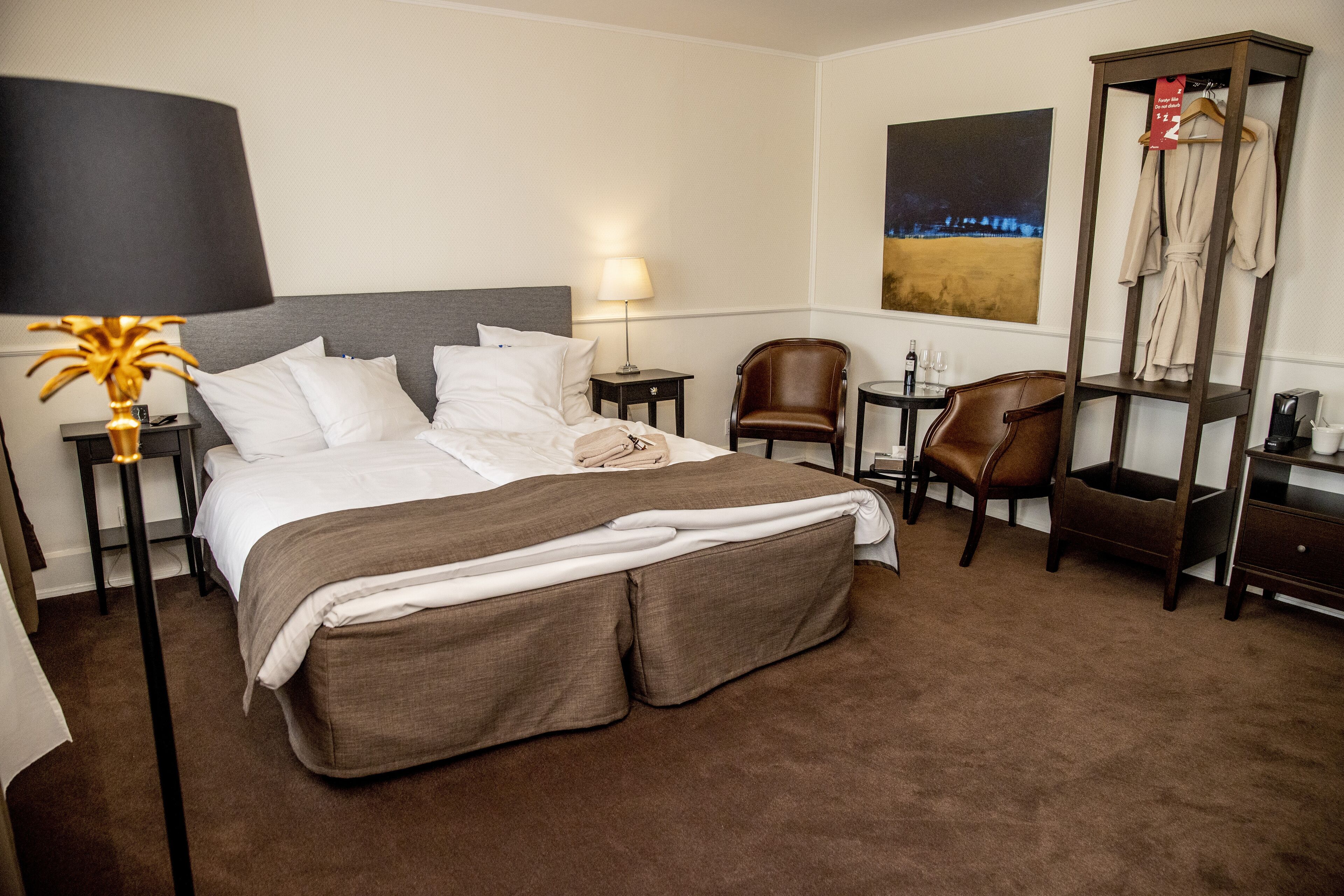 A hotel room with a large bed, a desk, a chair, a lamp, and a coat rack.