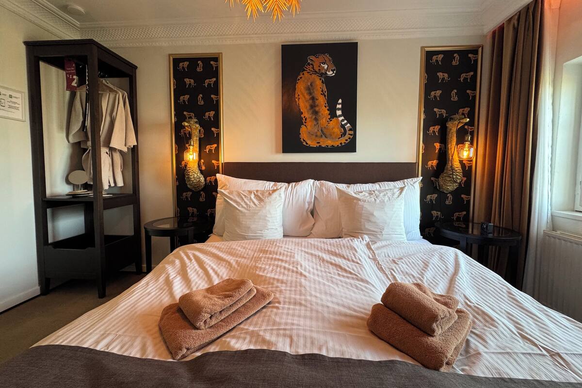A neatly made bed with two towels, a headboard, and a painting of a cheetah on the wall.