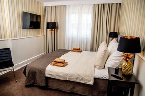 A hotel room with a bed, a TV mounted on the wall, a lamp, and a small table with a pineapple-shaped lamp. - Godstedlund B&B (Oster Ulslev)