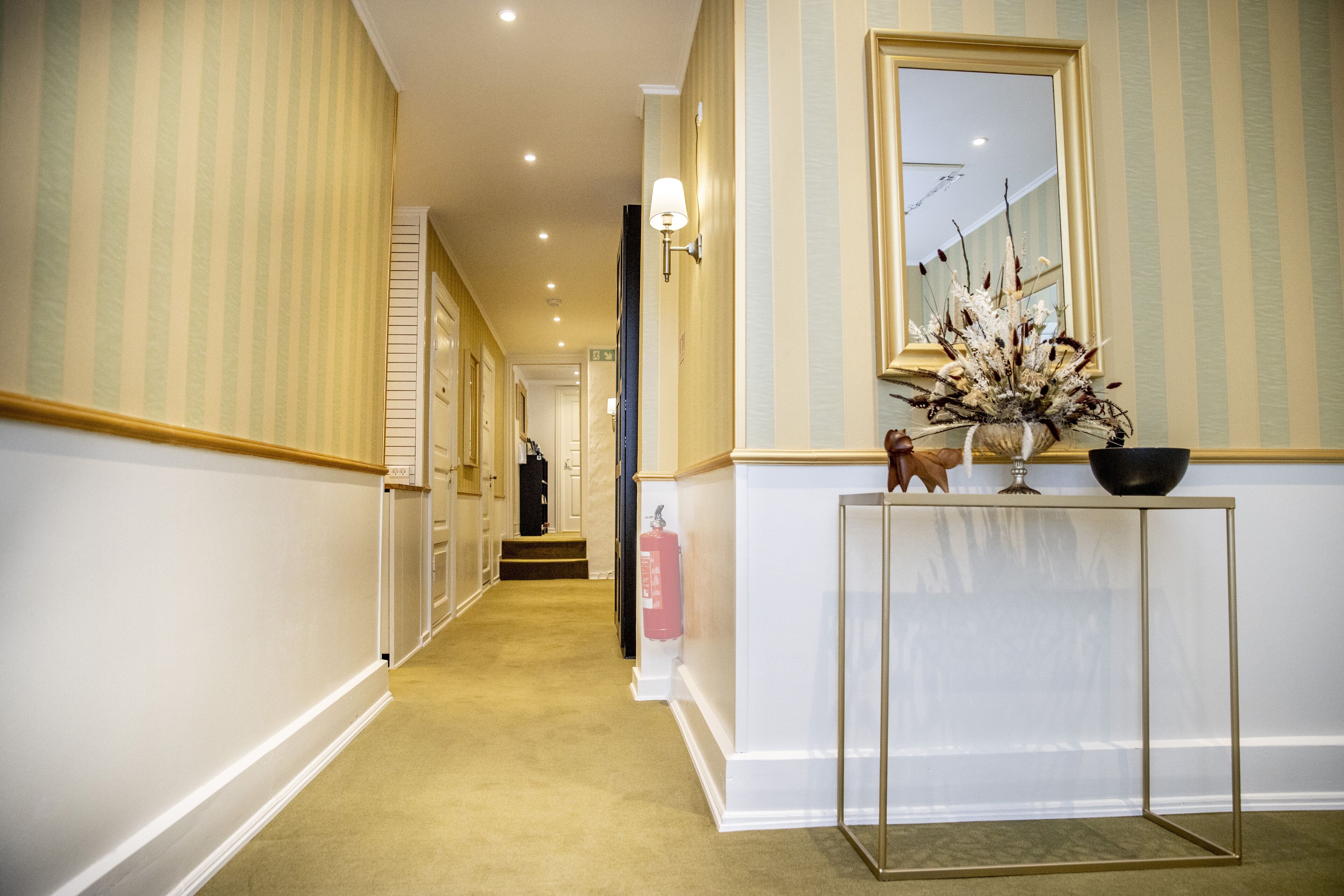 Un couloir d’hôtel avec un papier peint à rayures, une console sur laquelle sont posés un vase et un bol, et un extincteur.