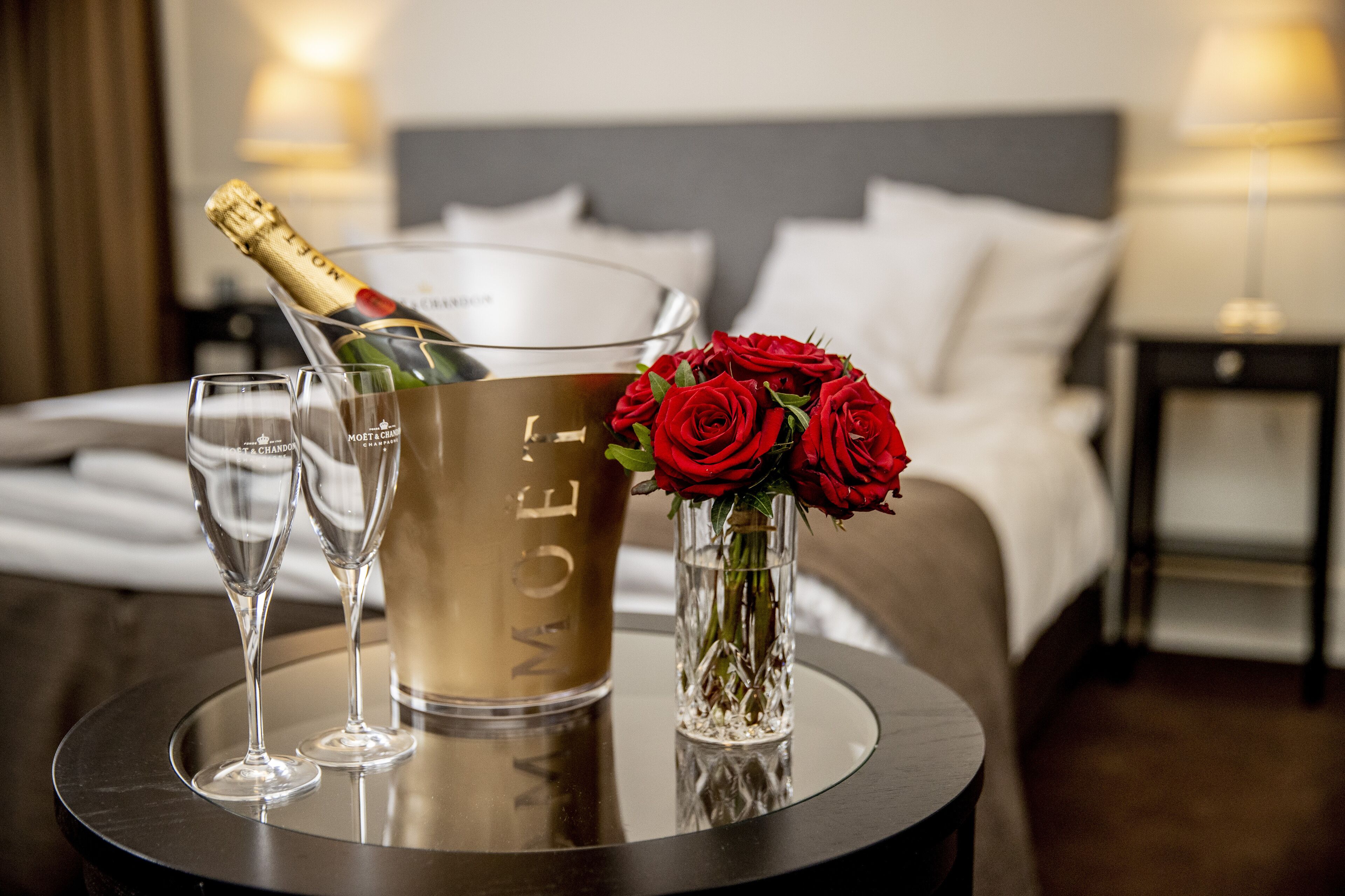 Une chambre d’hôtel avec un lit, une table de chevet sur laquelle se trouve une bouteille de champagne, deux flûtes à champagne et un vase rempli de roses rouges.