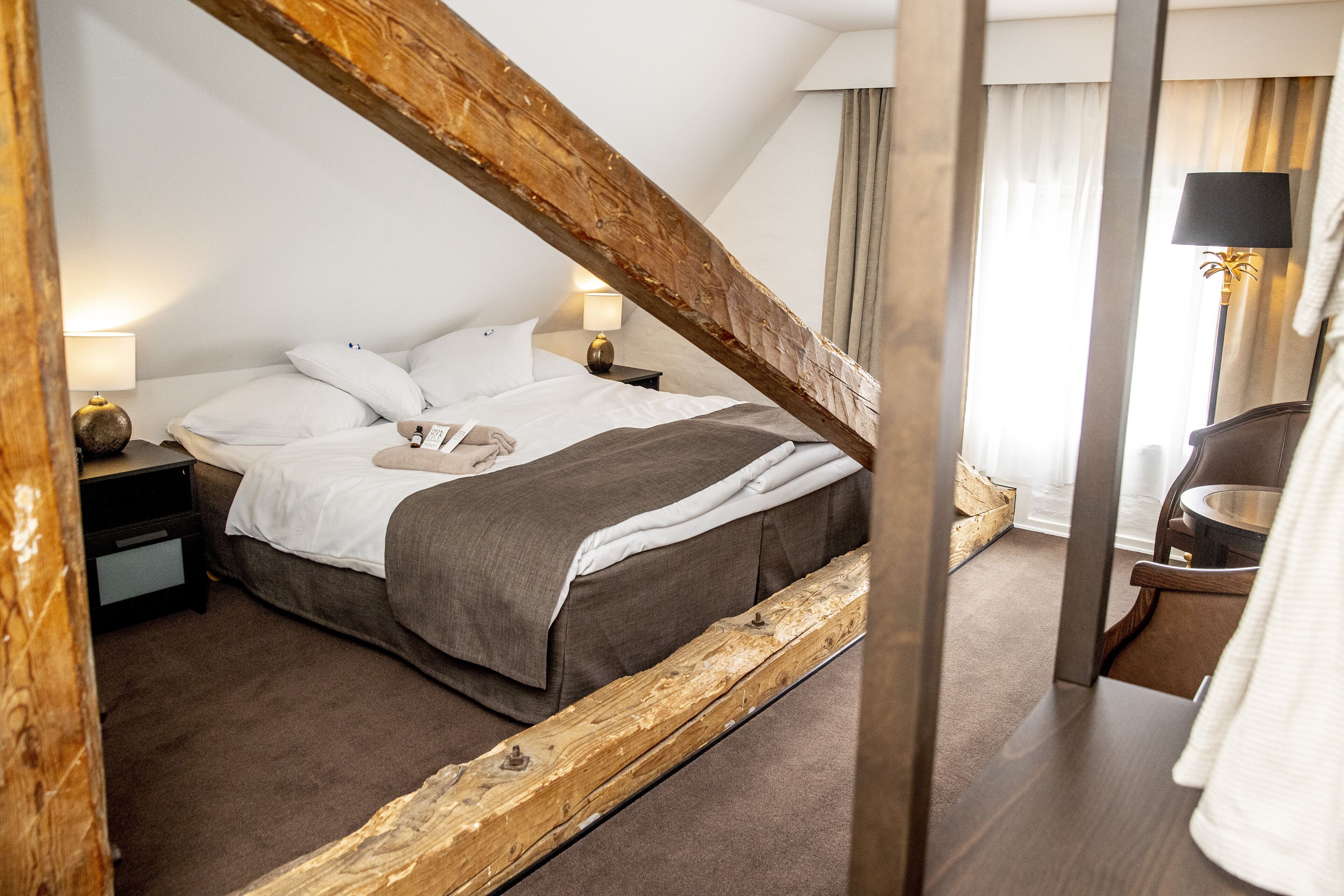 A bedroom with a wooden ceiling beam, a bed with white and gray bedding, a nightstand with a lamp, and a wooden chair.