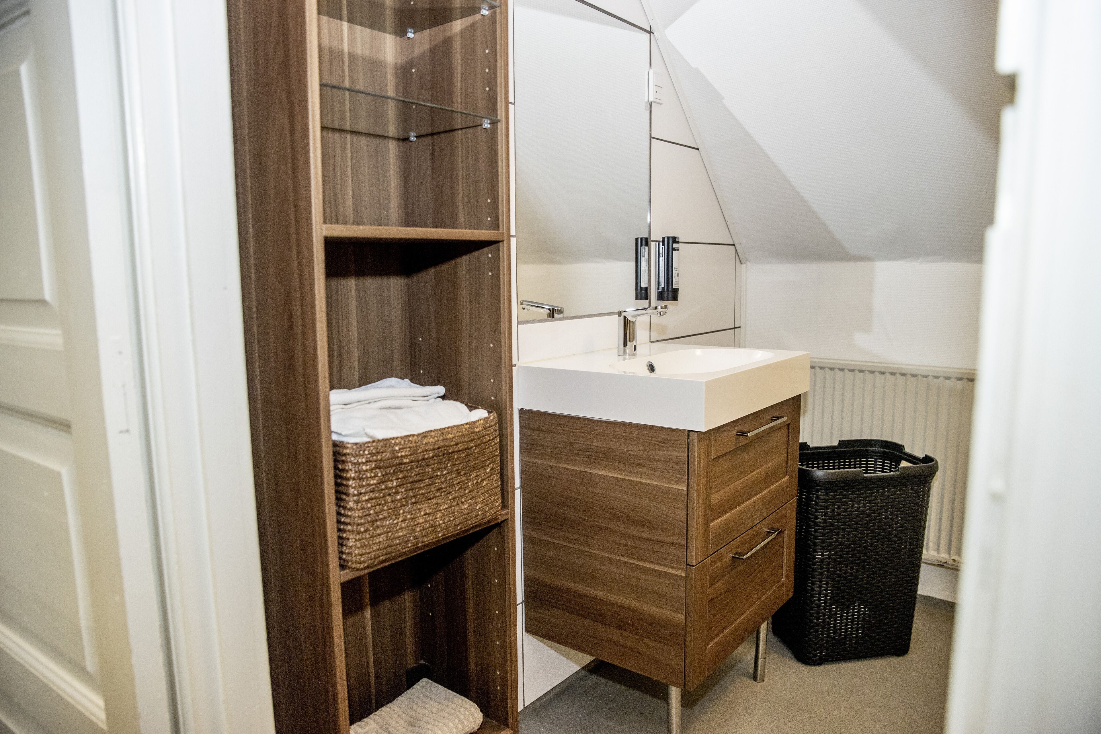 A bathroom with a wooden vanity, a white sink, and a mirror. There is a wicker basket on the shelf and a black trash bin.