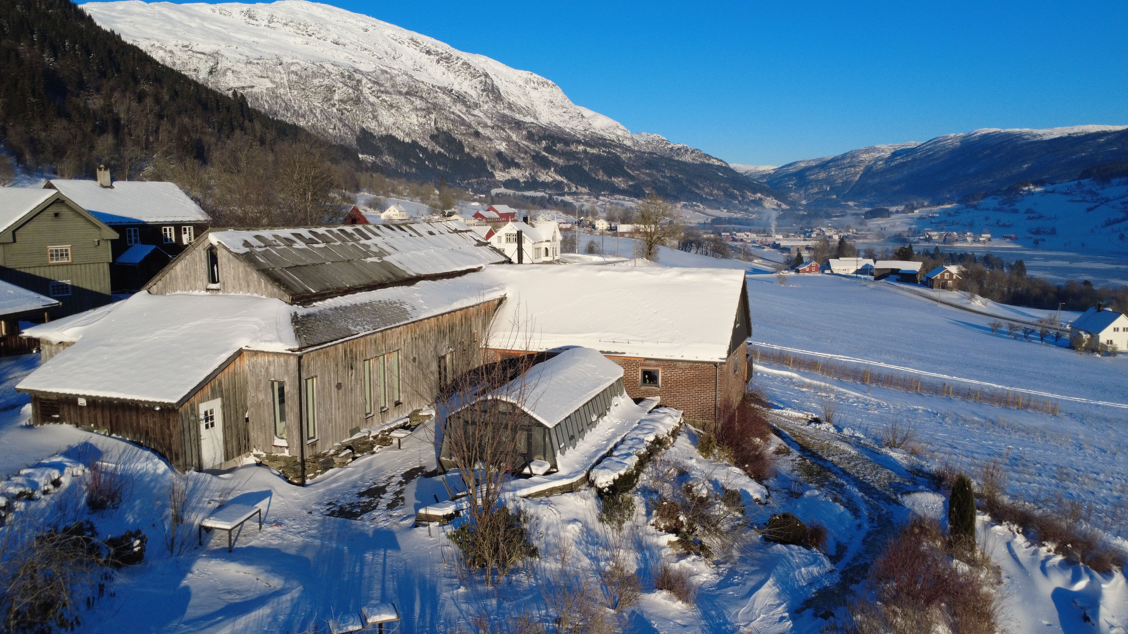 Paisaje rural nevado con edificios de madera tradicionales, cielo despejado y montañas al fondo.