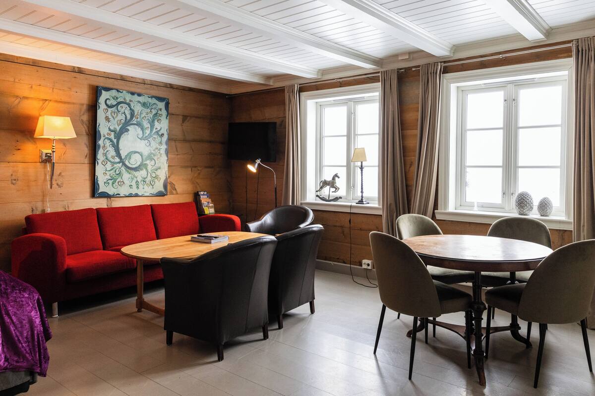 Een kamer met een houten plafond, een rode bank, een houten tafel met stoelen, een aan de muur bevestigde televisie en een raam met gordijnen.