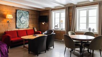 Una habitación con techo de madera, un sofá rojo, una mesa de madera con sillas, un televisor montado en la pared y un ventanal con cortinas.