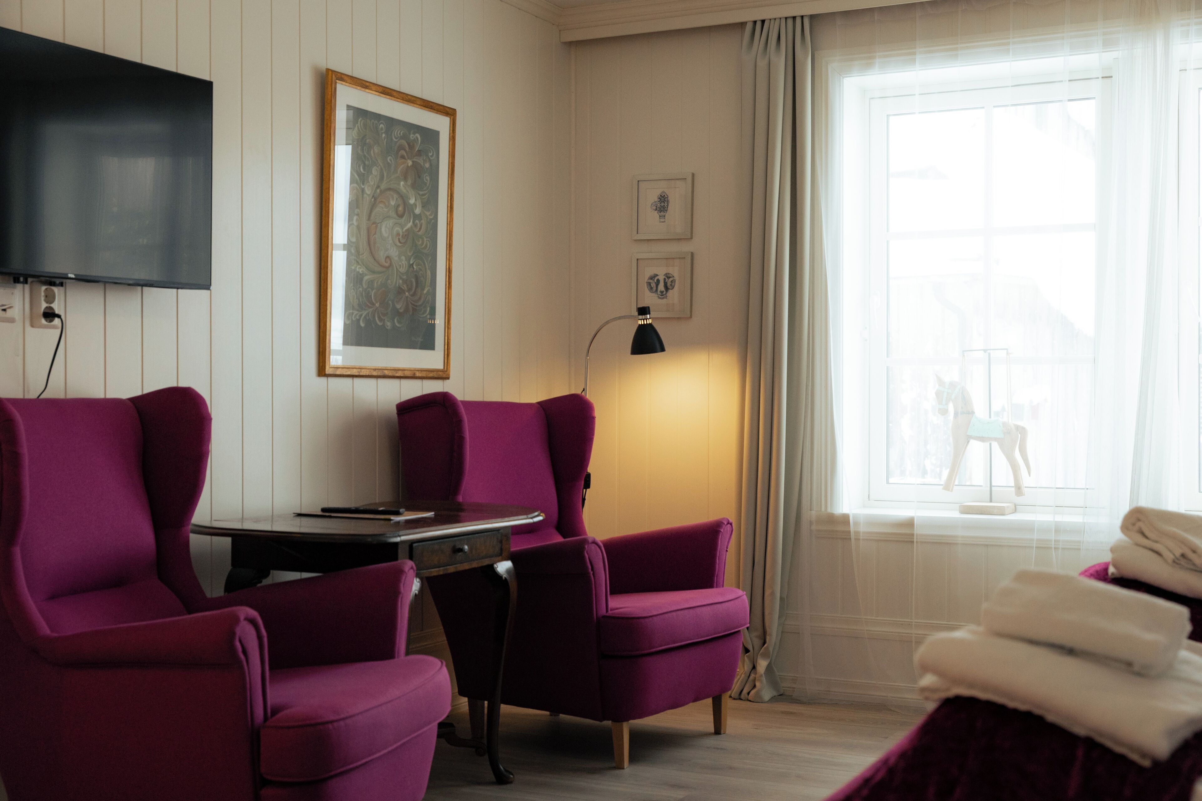 Een kamer met een aan de muur bevestigde televisie, een bureau met stoel, twee fauteuils, een lamp en een raam met gordijnen.
