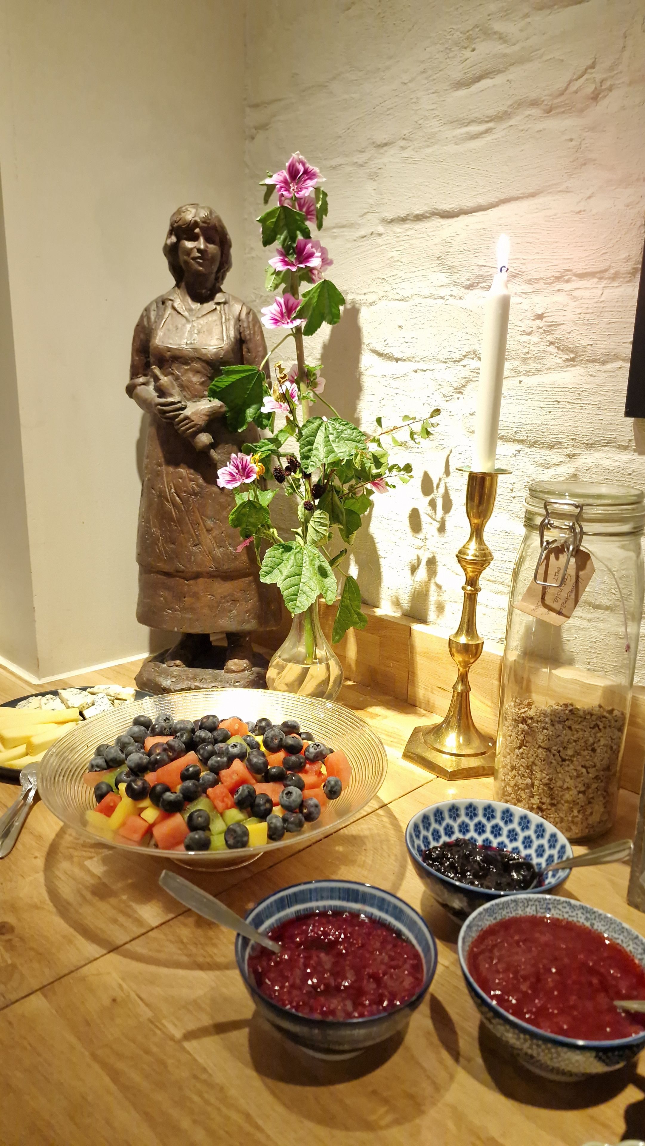 A table with a fruit salad, bowls of jam, a candle, a statue, and a vase with flowers.