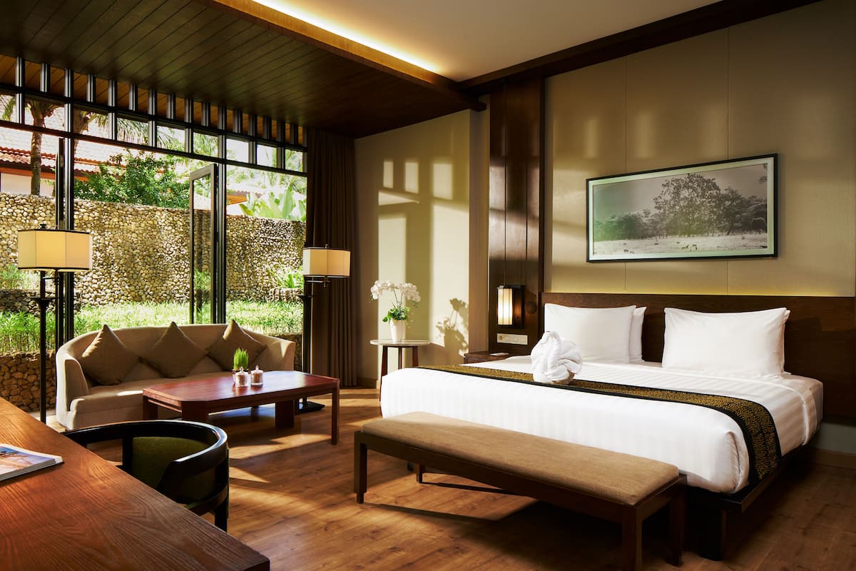 Habitación de hotel con cama, sofá, mesita de centro y escritorio. Se ve vegetación a través de la ventana.
