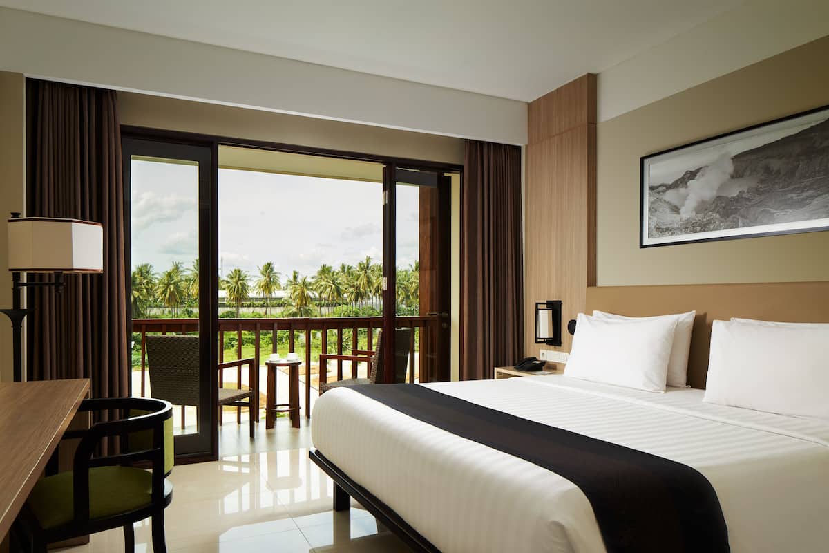 Habitación de hotel con una cama grande, un escritorio y una silla. Tiene un balcón con vistas a palmeras y una montaña.