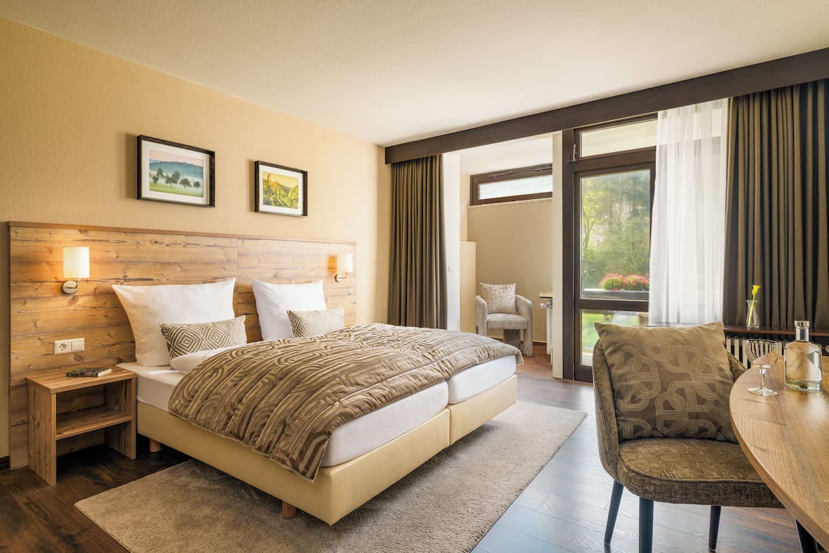 Hotelkamer met een groot bed, een houten hoofdeinde, een nachtkastje, een stoel, een tafel en uitzicht op de omgeving door een groot raam.