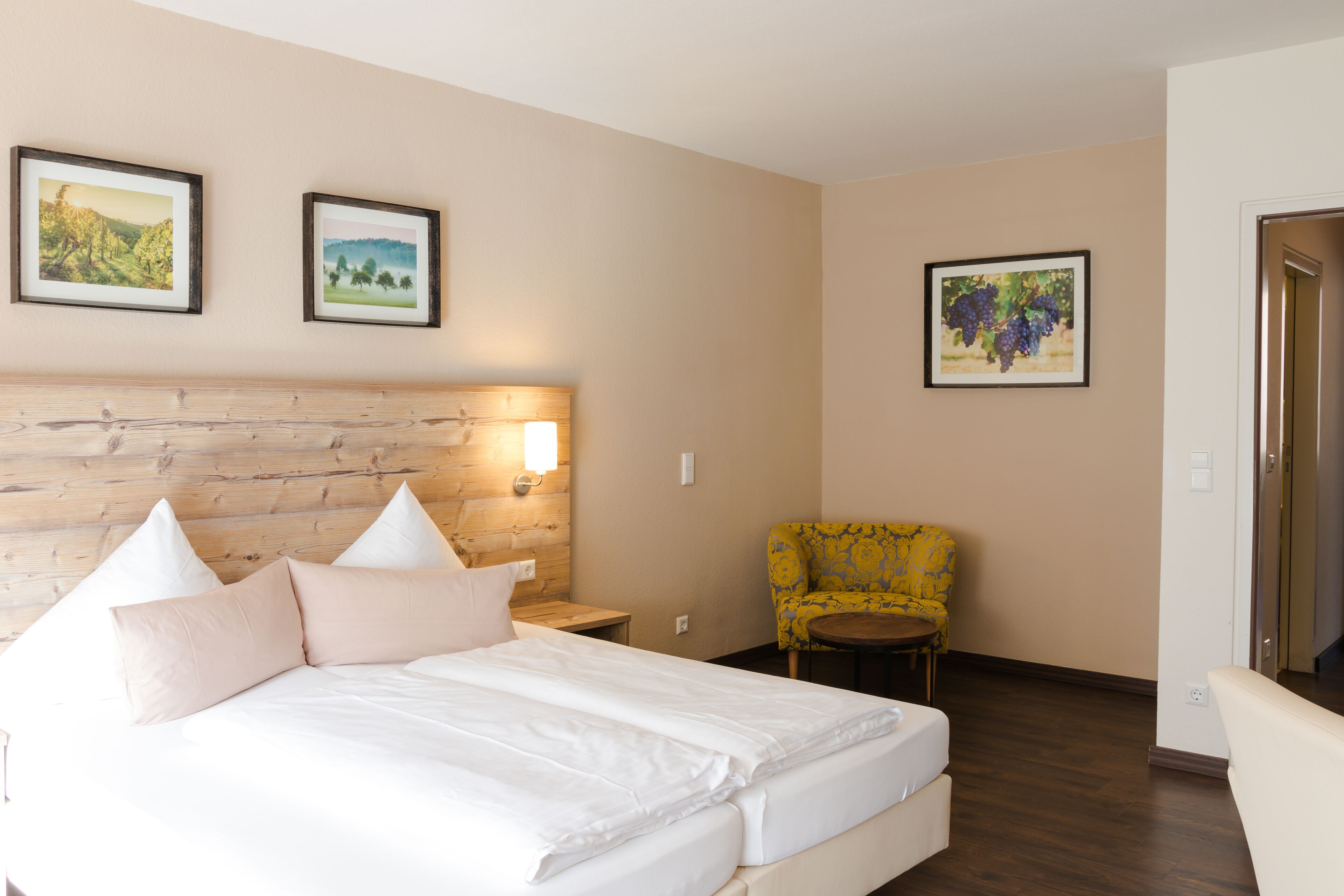Ein Hotelzimmer mit einem Holz-Kopfteil, einem Bett mit weißer Bettwäsche, einem kleinen runden Tisch, einem gelben Sessel und zwei gerahmten Bildern an der Wand.