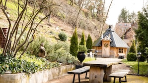 Une cabane en bois avec un toit en pente, entourée d’un jardin agrémenté d’un banc en bois et d’un grand bac à fleurs.