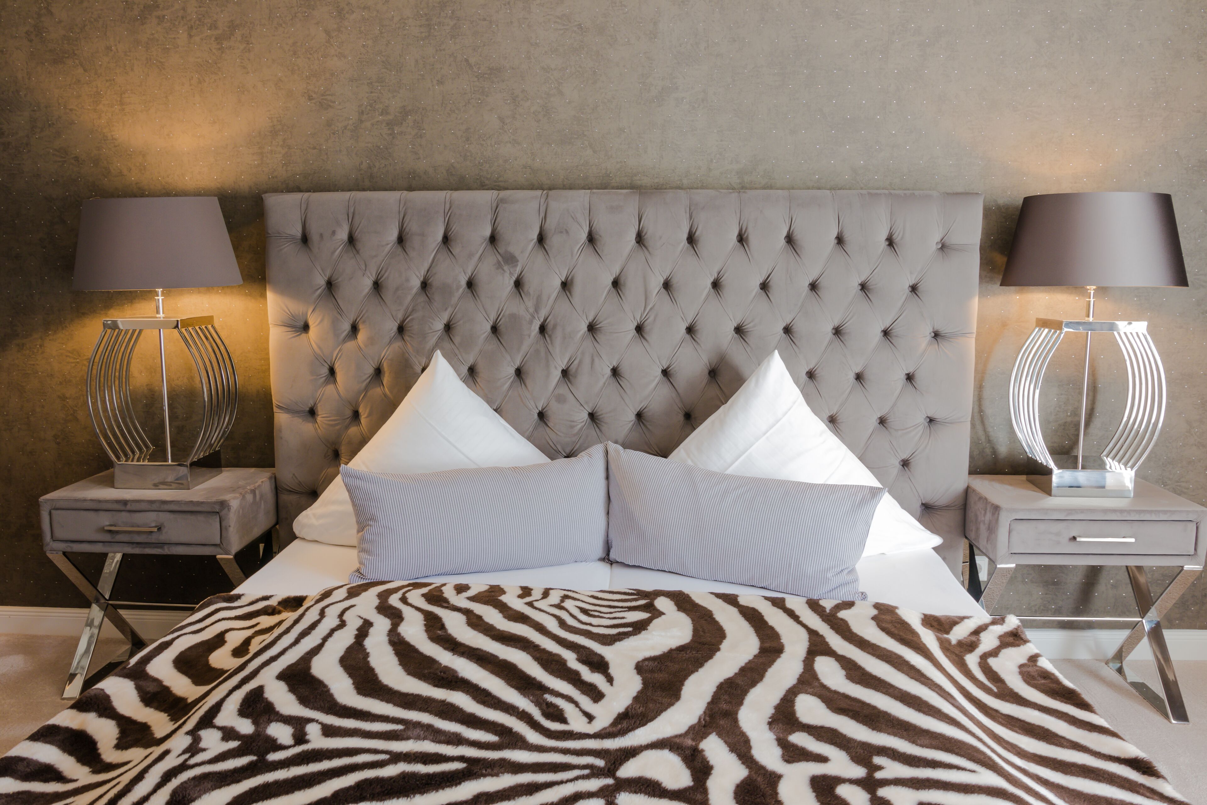 A bedroom with a tufted headboard, two bedside tables with lamps, a zebra-patterned bedspread, and two pillows.