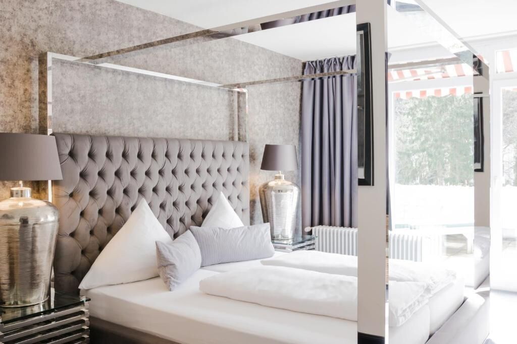 Camera d'hotel moderna con testiera trapuntata, un letto grande con lenzuola bianche, due abat-jour e vista sull'esterno attraverso una grande finestra.