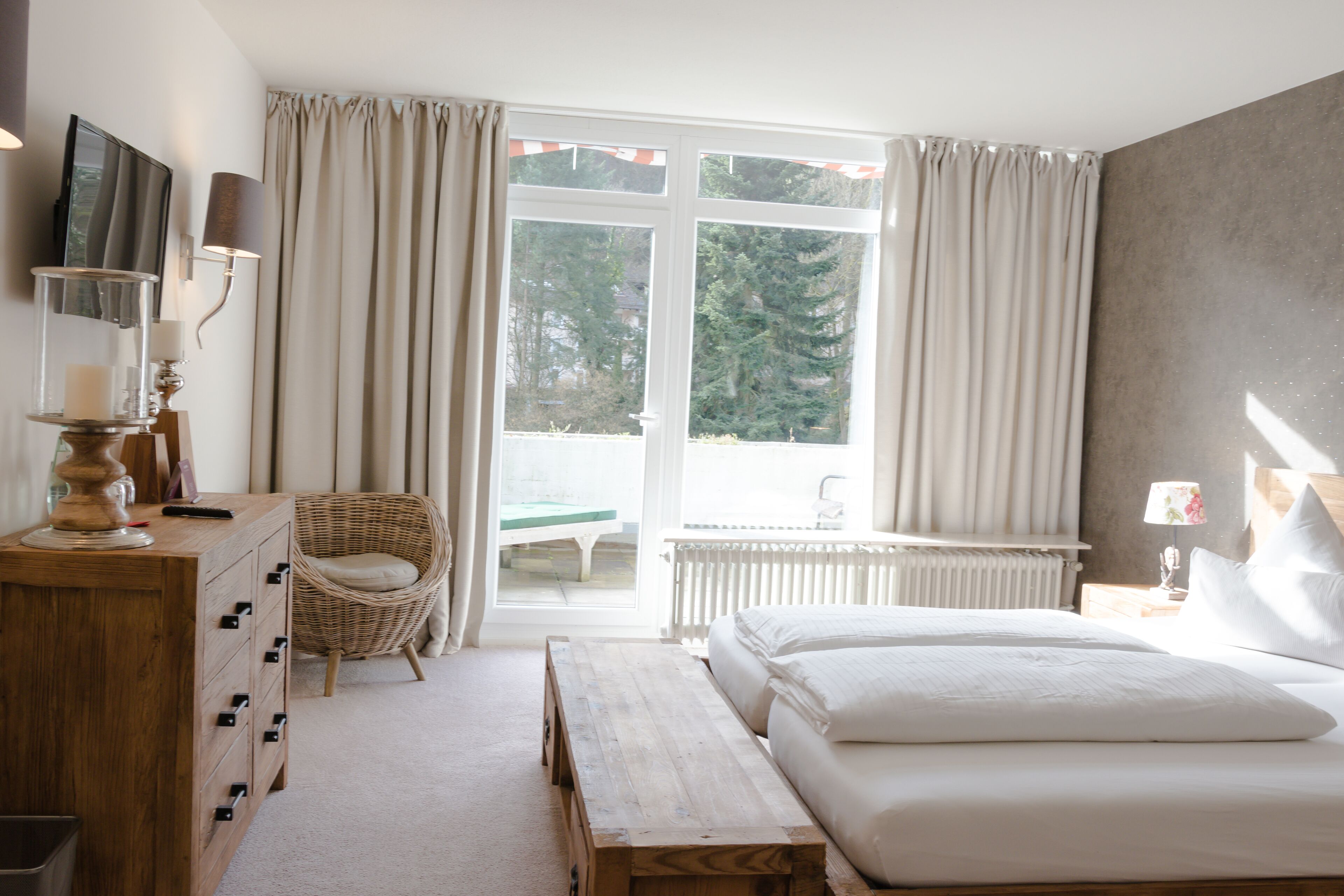 Un dormitorio con una cama, un aparador de madera, una silla de mimbre, un banco, una ventana con cortinas y vistas a árboles fuera.