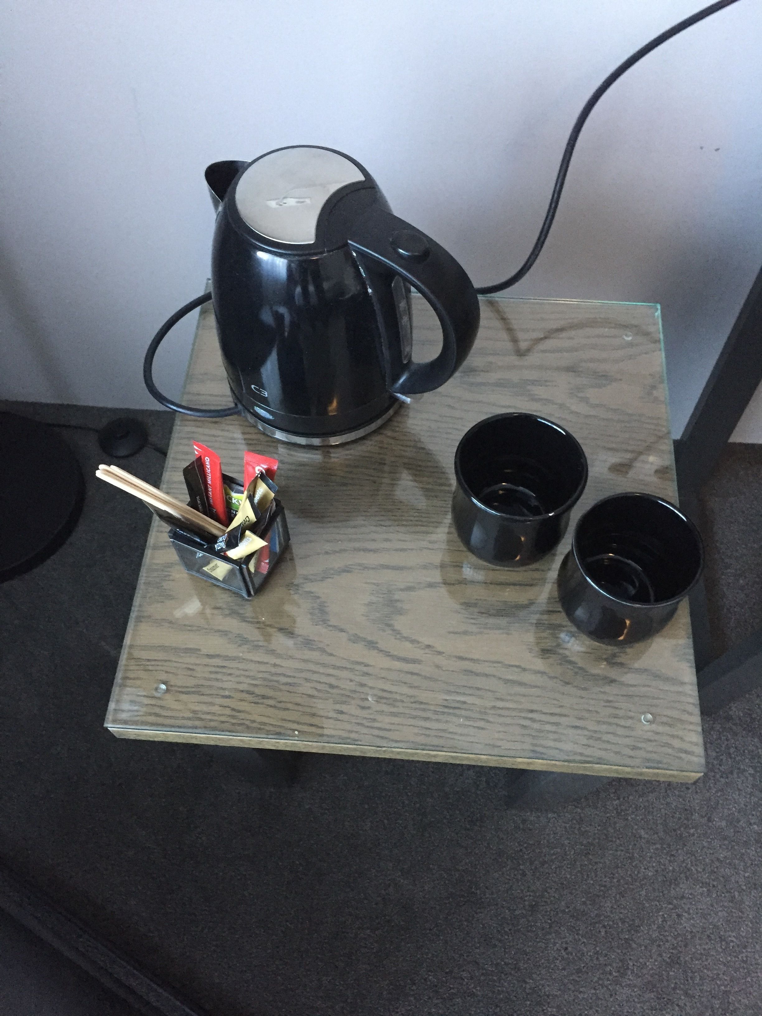 A wooden table with a black electric kettle, two black cups, and a set of chopsticks.