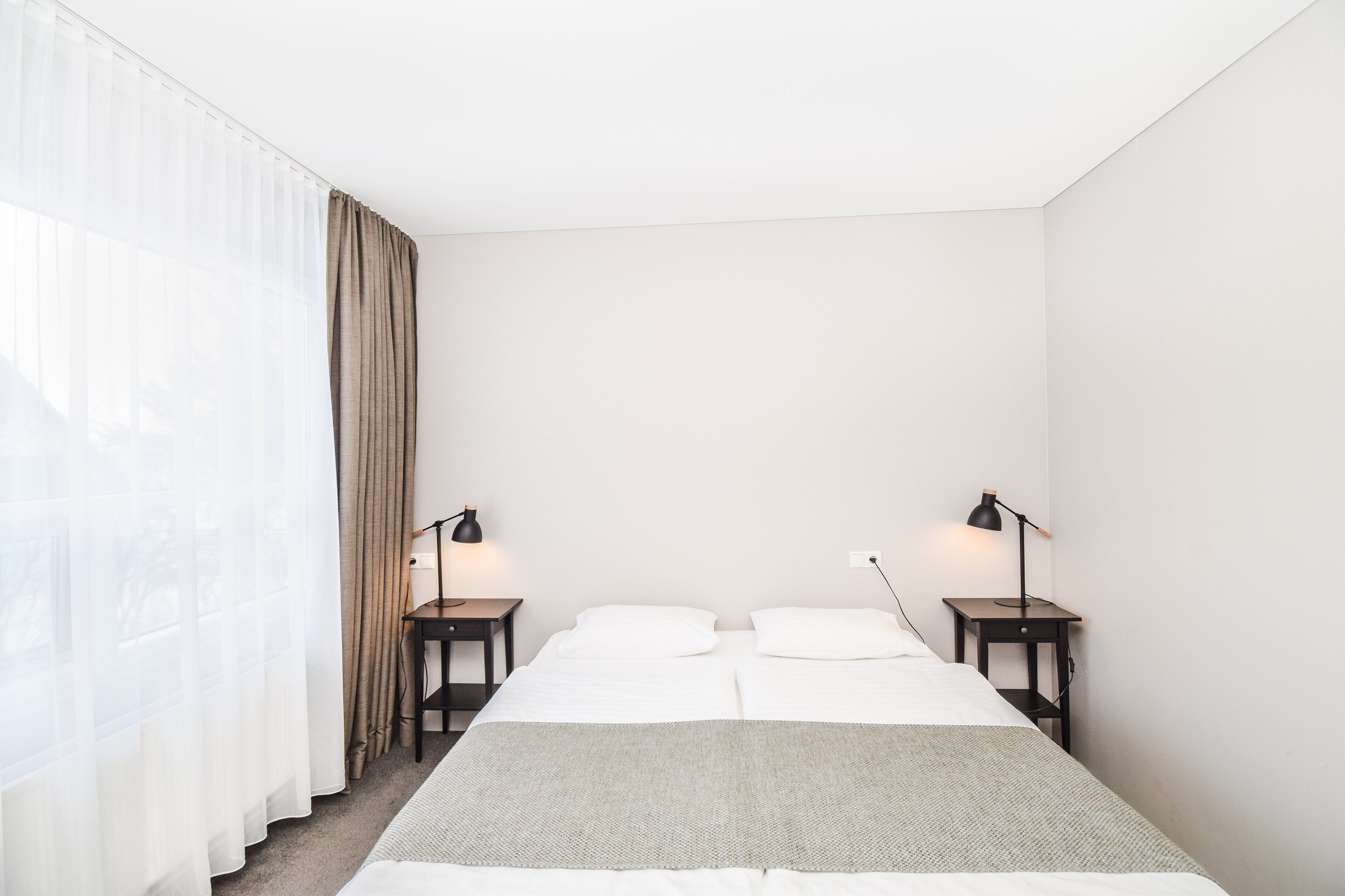 Habitación de hotel minimalista con cama, dos mesitas de noche y ventana con cortinas de gasa.