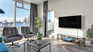 Ein modernes Wohnzimmer mit großem Fernseher, bequemen Sitzmöglichkeiten, einem Couchtisch und Blick auf die Stadt.