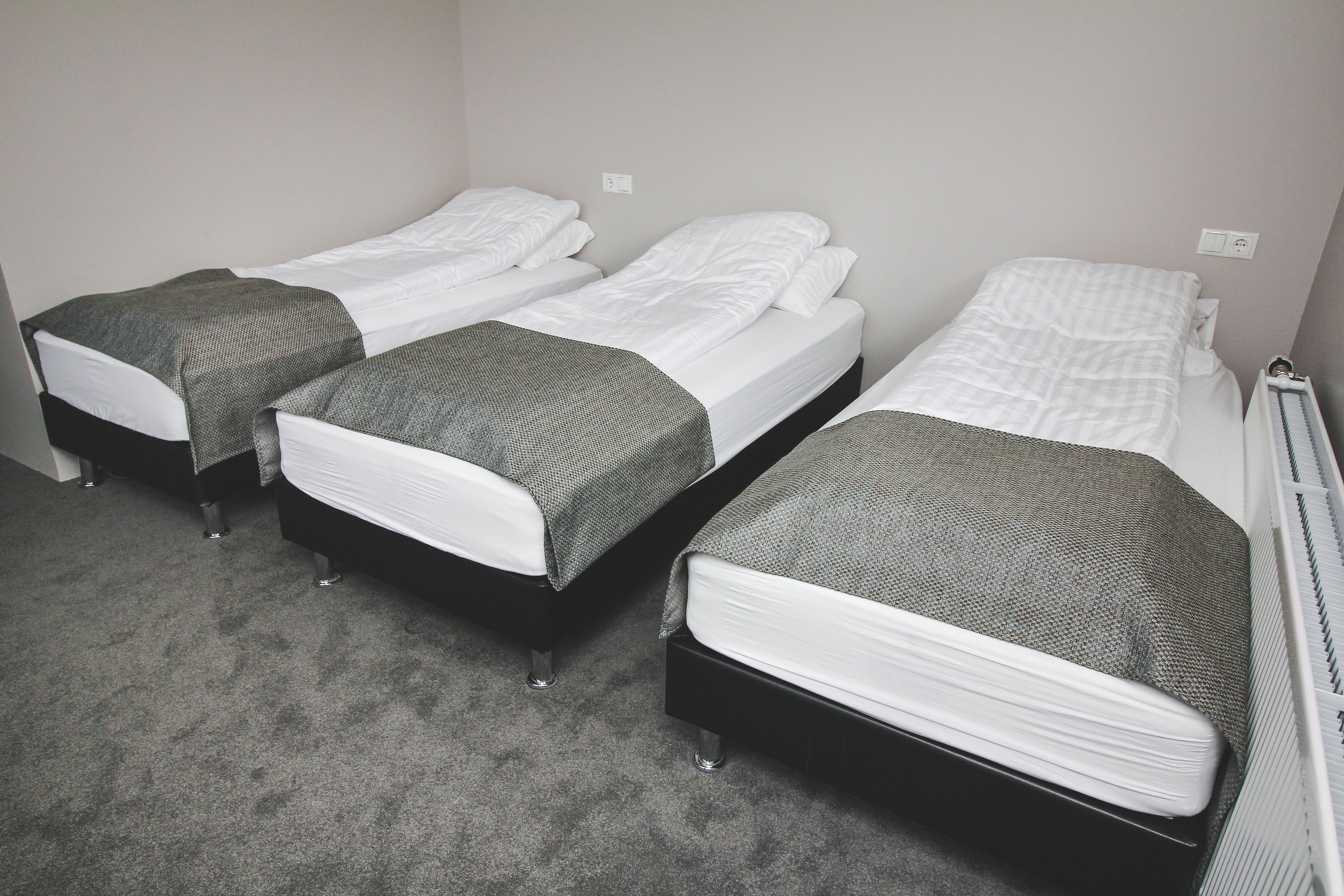 Una habitación con tres camas, cada una con ropa de cama blanca y mantas grises. El piso está alfombrado y las paredes son blancas.