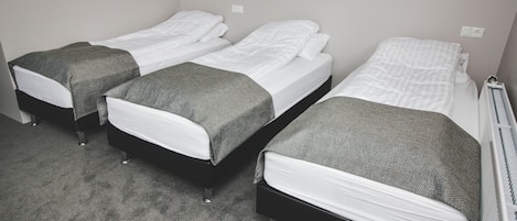 Una habitación con tres camas, cada una con ropa de cama blanca y mantas grises. El piso está alfombrado y las paredes son blancas.