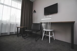 Una habitación con escritorio, dos sillas, televisor montado en la pared y ventana con cortinas.