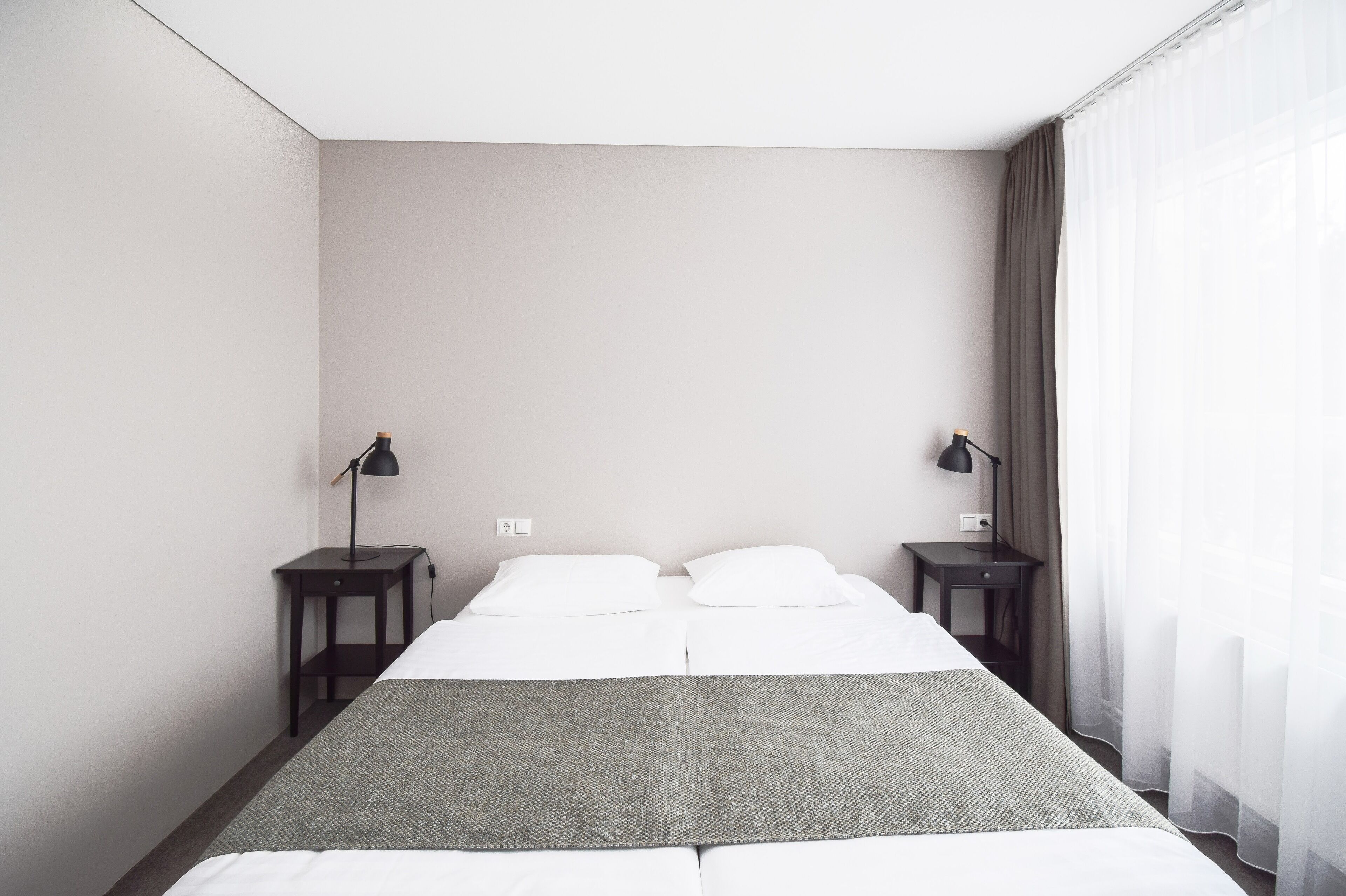 A hotel room with a bed, two bedside tables, two lamps, a window with curtains, and a neutral color scheme.