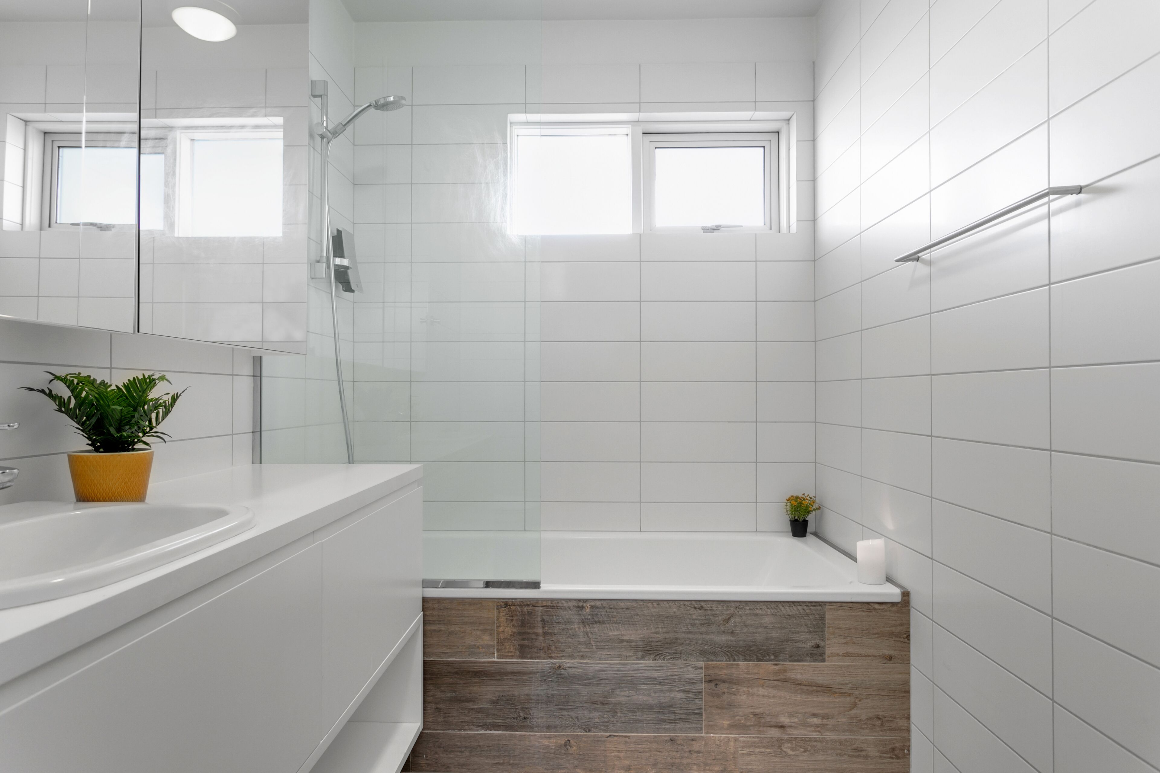 A modern bathroom with a white sink, mirror, and bathtub. There is a window above the bathtub and a potted plant on the counter.