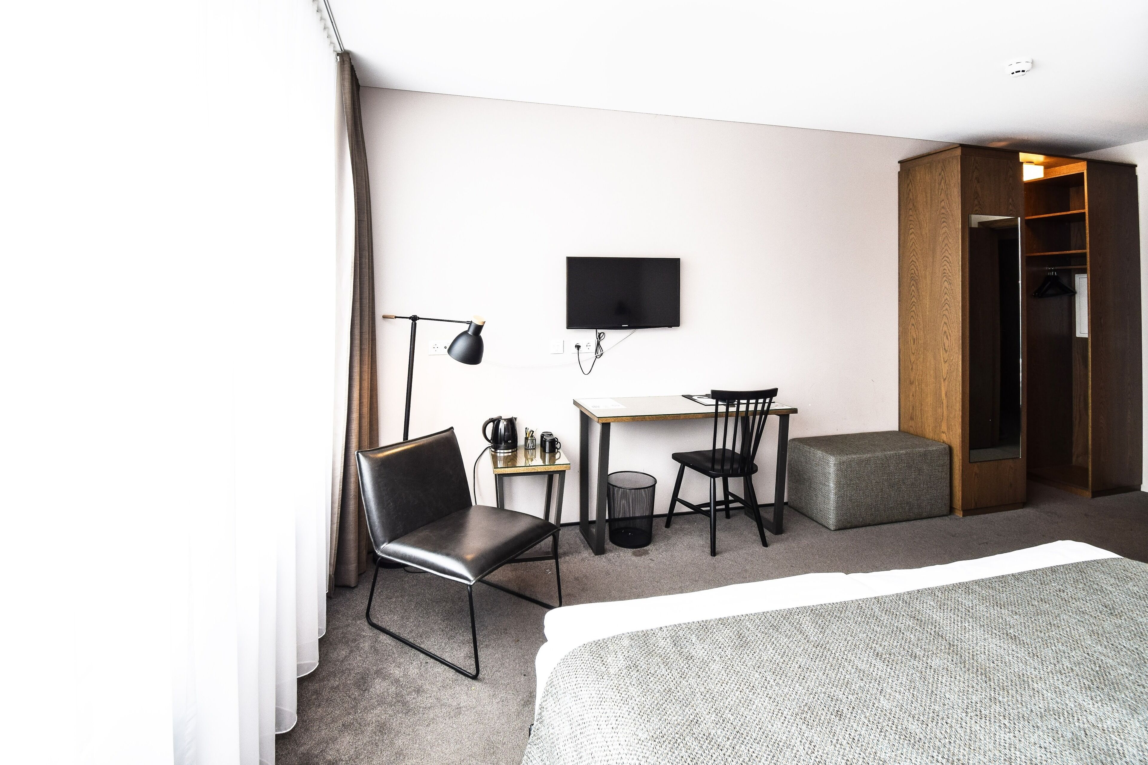 A hotel room with a bed, a desk with a chair, a TV, a wardrobe, and a small table with a lamp.