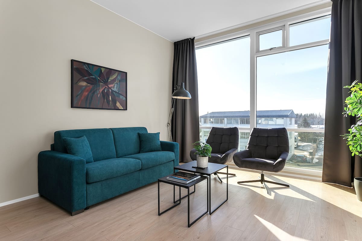 Una sala de estar moderna con un sofá azul verdoso, un sillón negro, una mesa de centro y un ventanal con vistas a edificios.