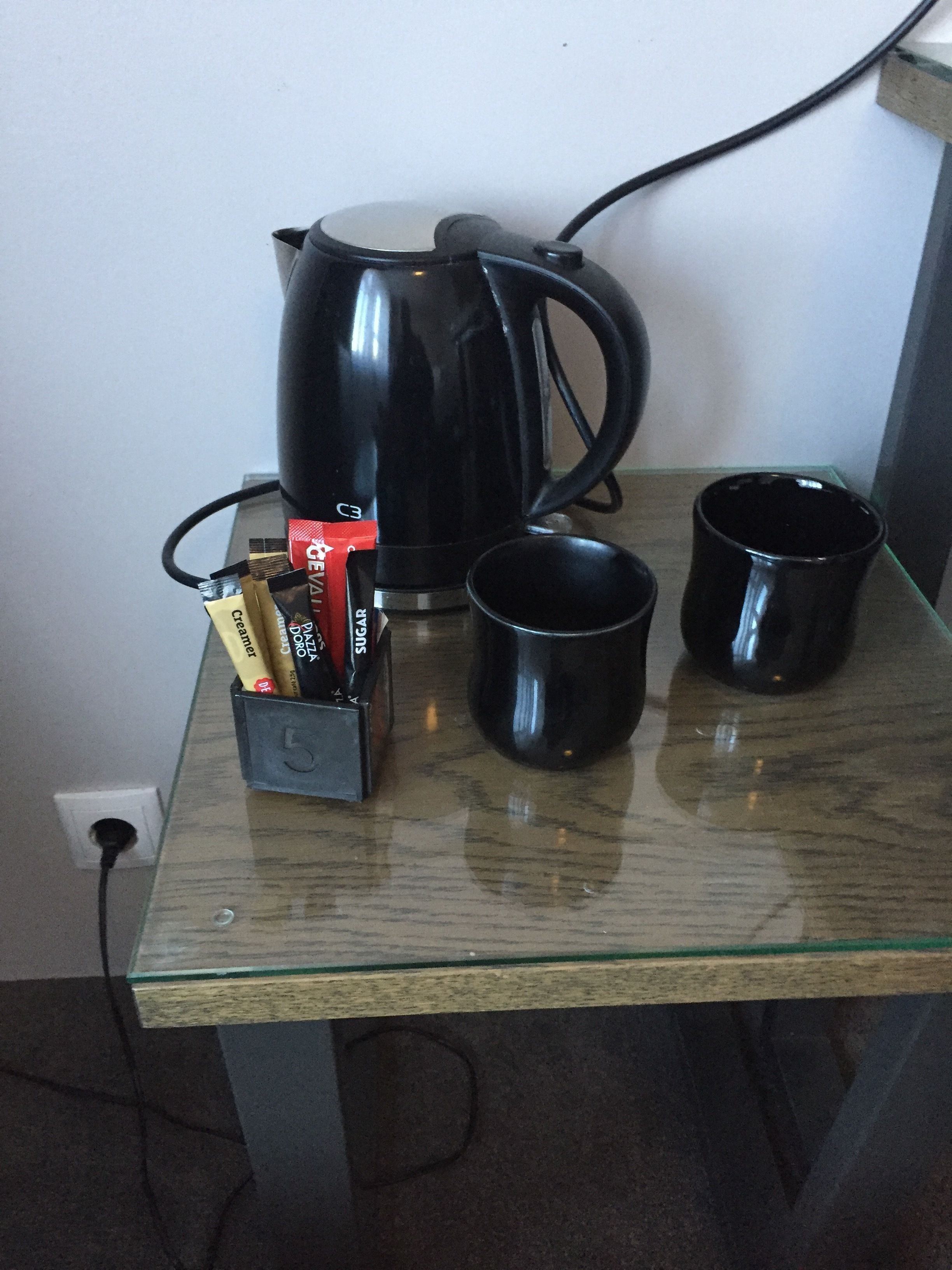 A glass table with a black coffee maker, two black mugs, and a box of coffee pods.