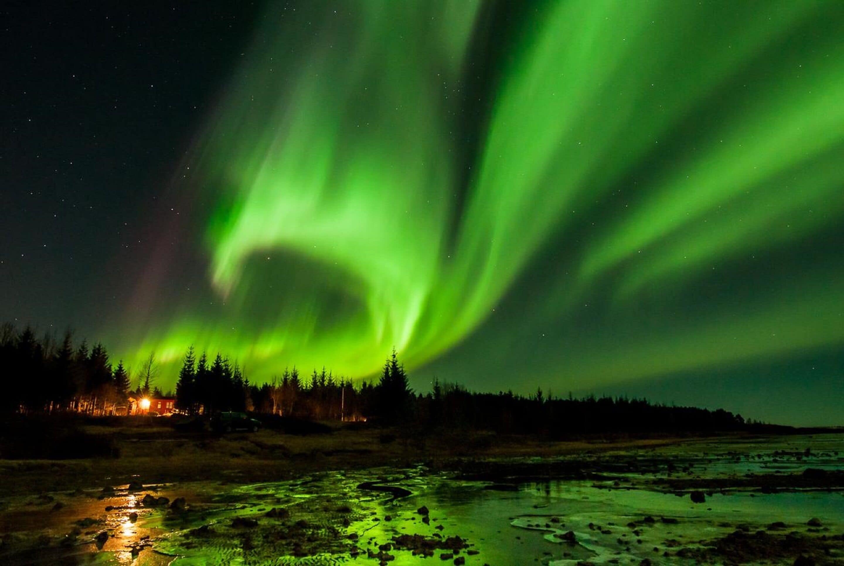 Una escena nocturna con auroras boreales sobre un paisaje que incluye un edificio y árboles.