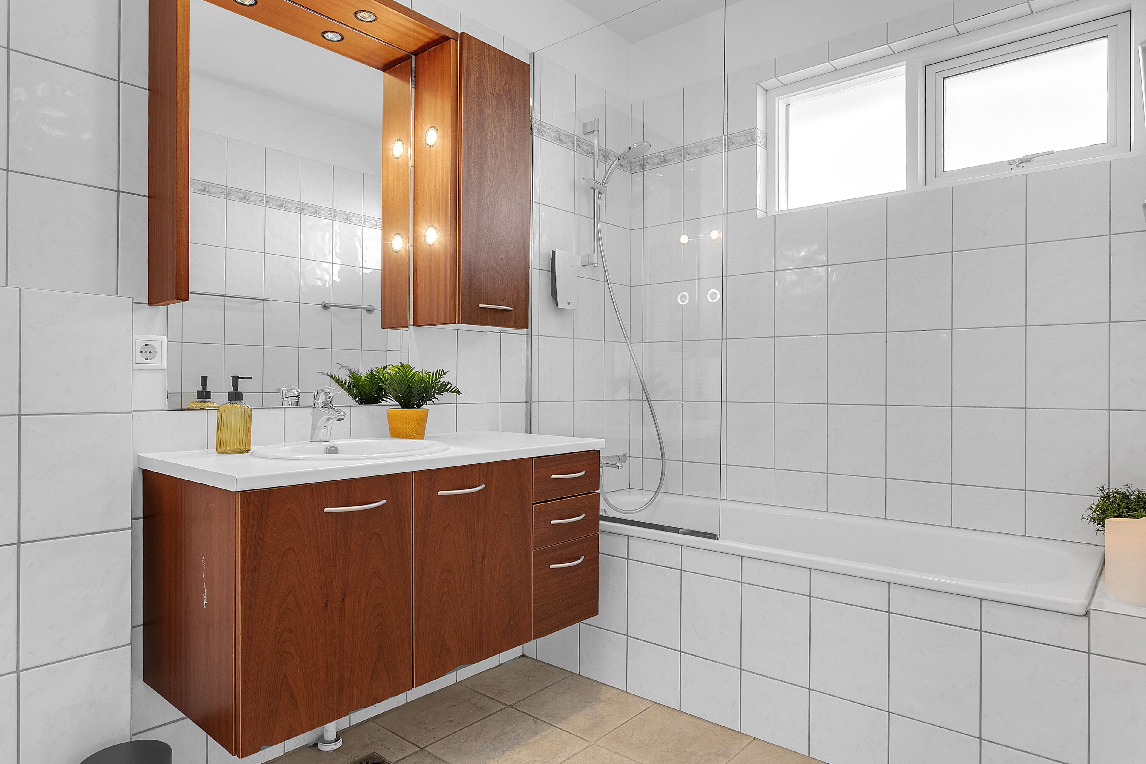 A bathroom with a wooden vanity, white tiles, a shower, and a window.