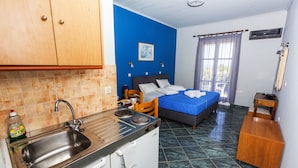 A compact hotel room with a kitchenette, a bed with blue bedding, a wooden chair, a small wooden table, and a window with curtains. - Anastasia Studios (Kefalonia)