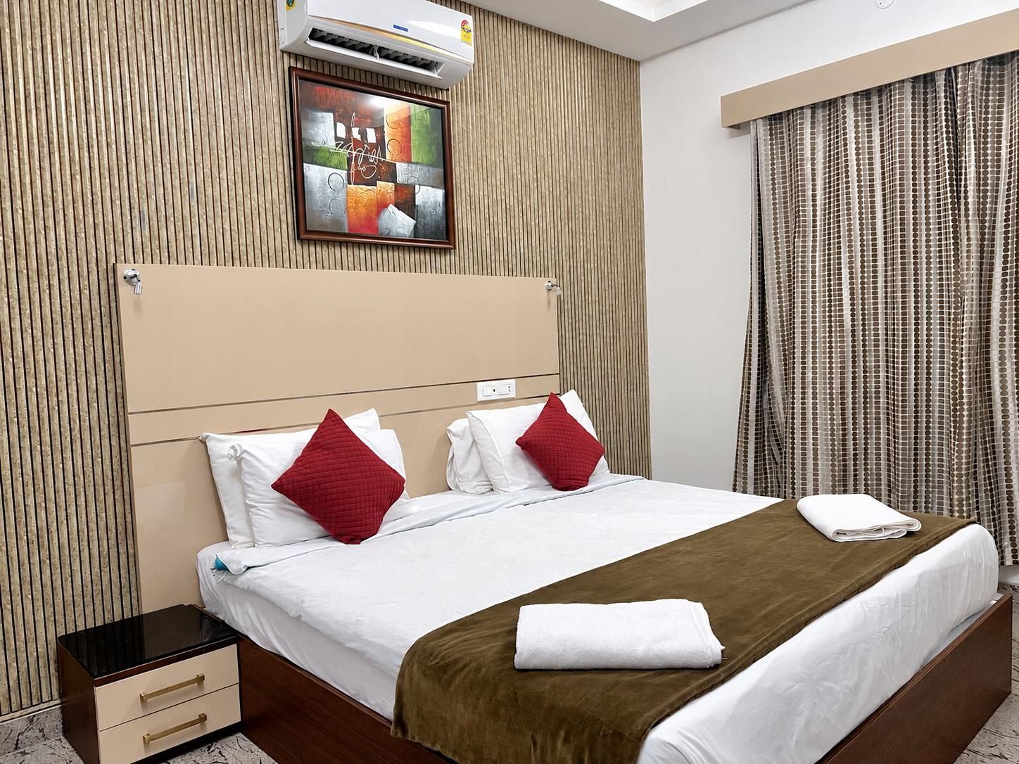 A hotel room with a bed, a nightstand, a wall-mounted air conditioner, and a window with curtains.