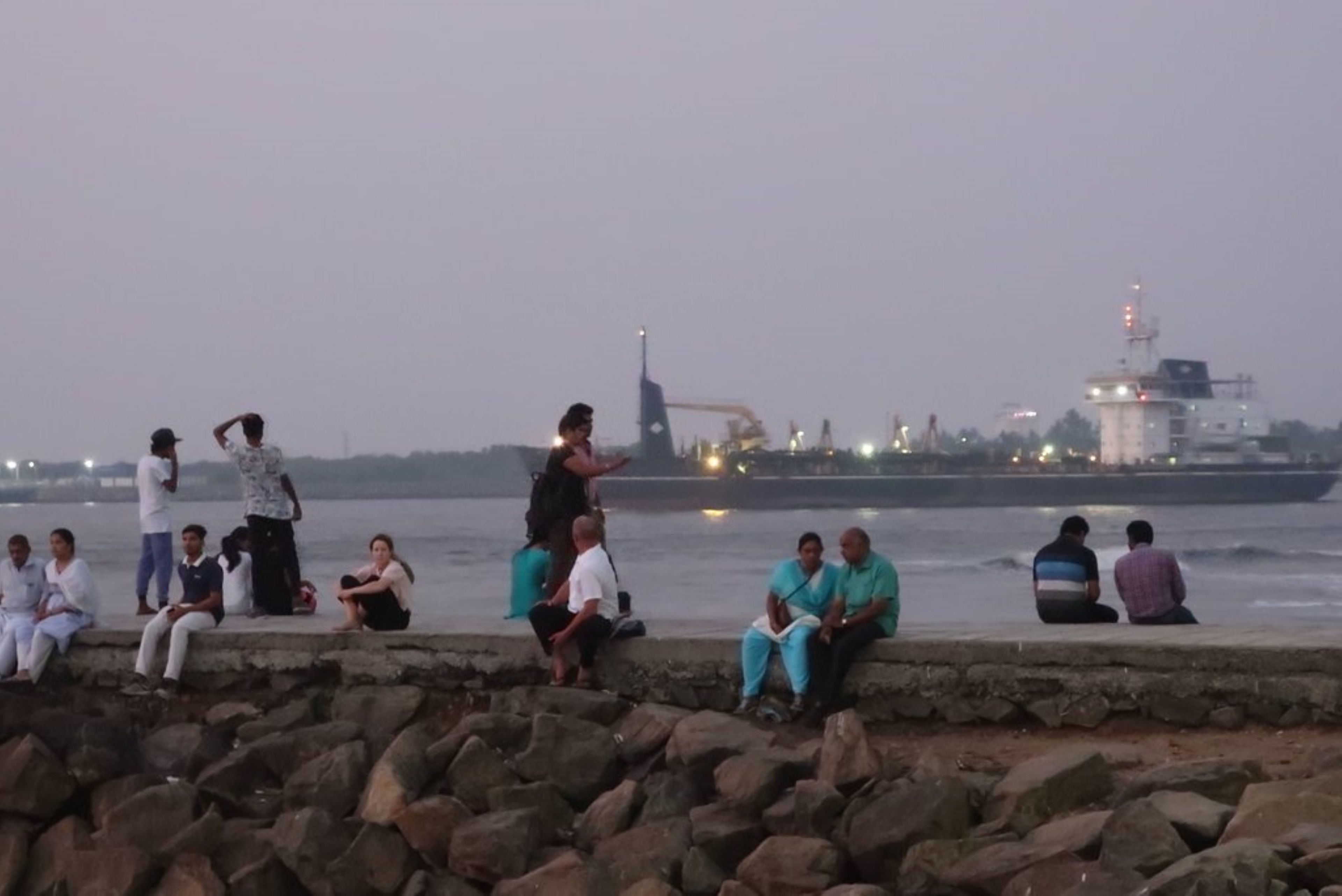 A group of people sitting and standing by a waterfront, with industrial structures in the background.