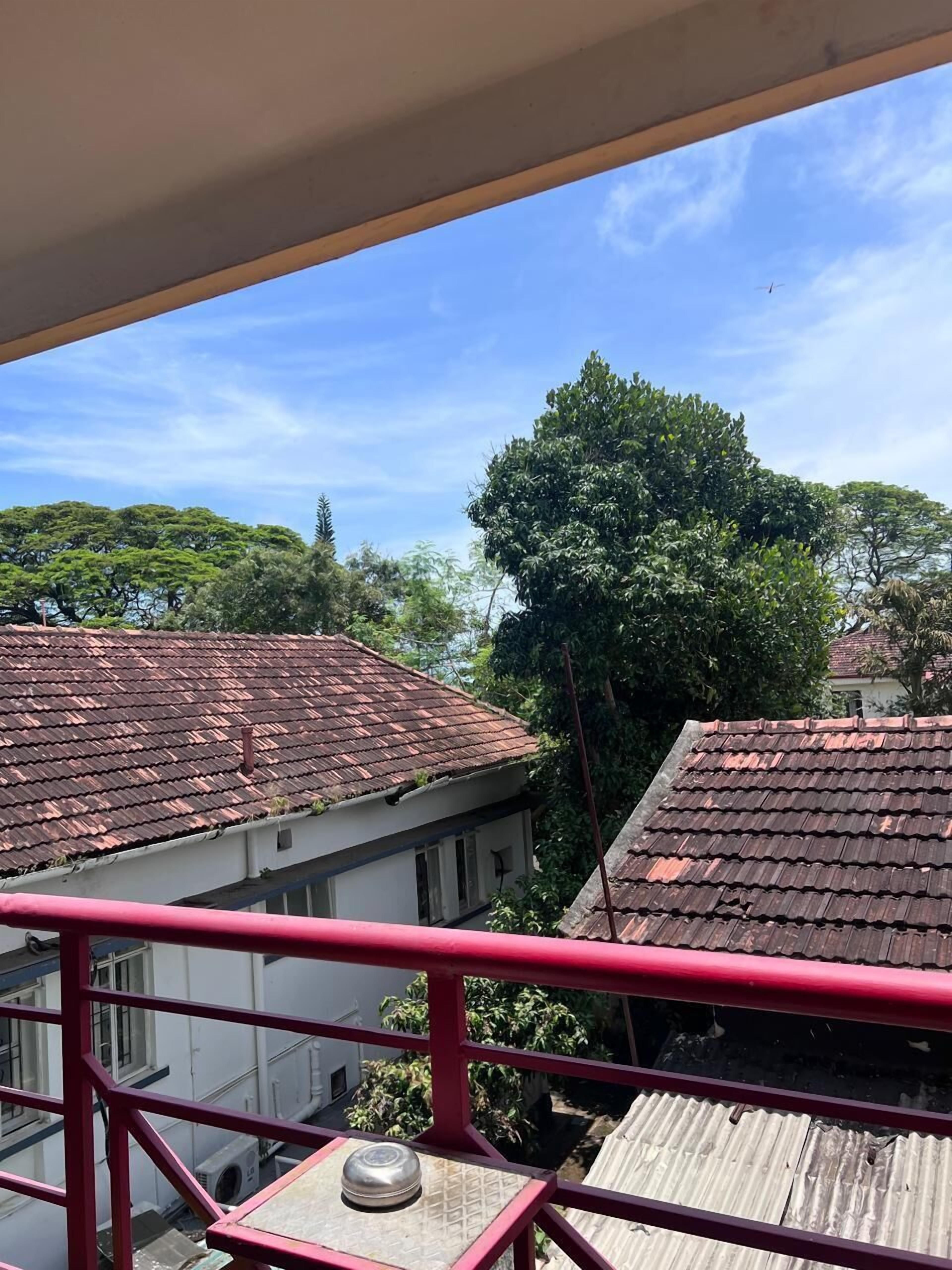 A view from a balcony with red railings, overlooking a residential area with houses that have red-tiled roofs and lush greenery.