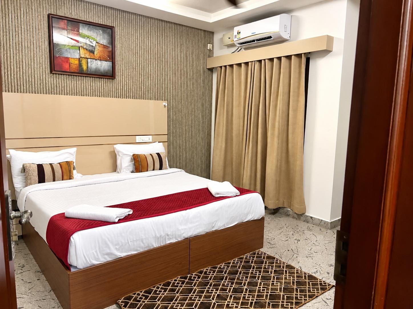 A hotel room with a large bed, a headboard, a wall-mounted air conditioner, and a painting.