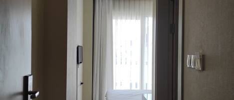 Habitación de hotel con cama, mesita de noche, ventana con cortinas, puerta y un interruptor de luz en la pared.