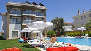 Zona de piscina con sillones de descanso y sombrillas, un edificio con balcones y cielo despejado.