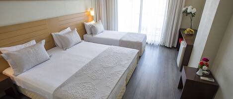 Habitación de hotel con dos camas, una mesita de noche, un escritorio, una ventana con cortinas y una pequeña mesa con un florero.