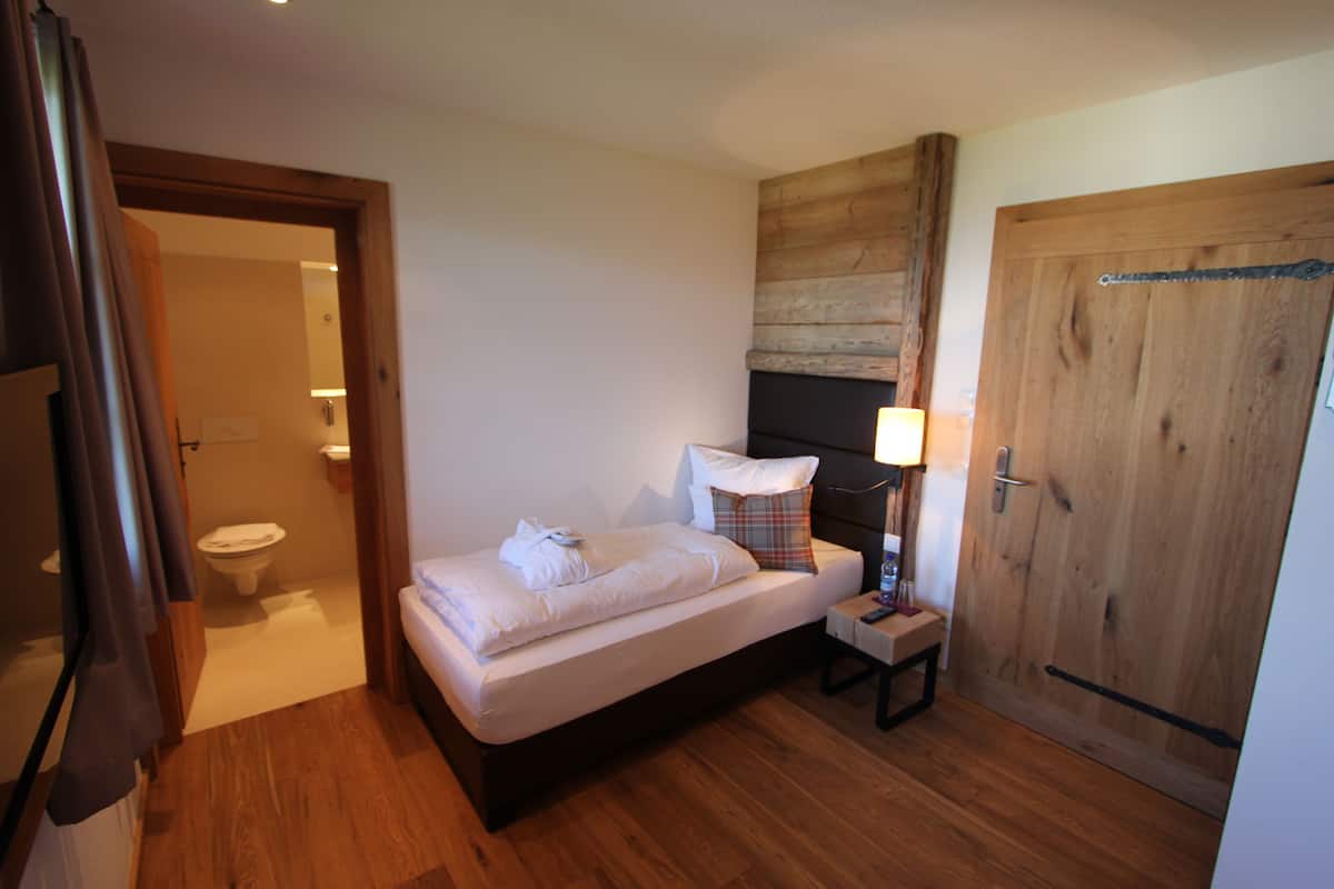 Un dormitorio pequeño y acogedor con piso de madera, una cama con sábanas blancas, una mesita de noche con una lámpara y una puerta de madera.