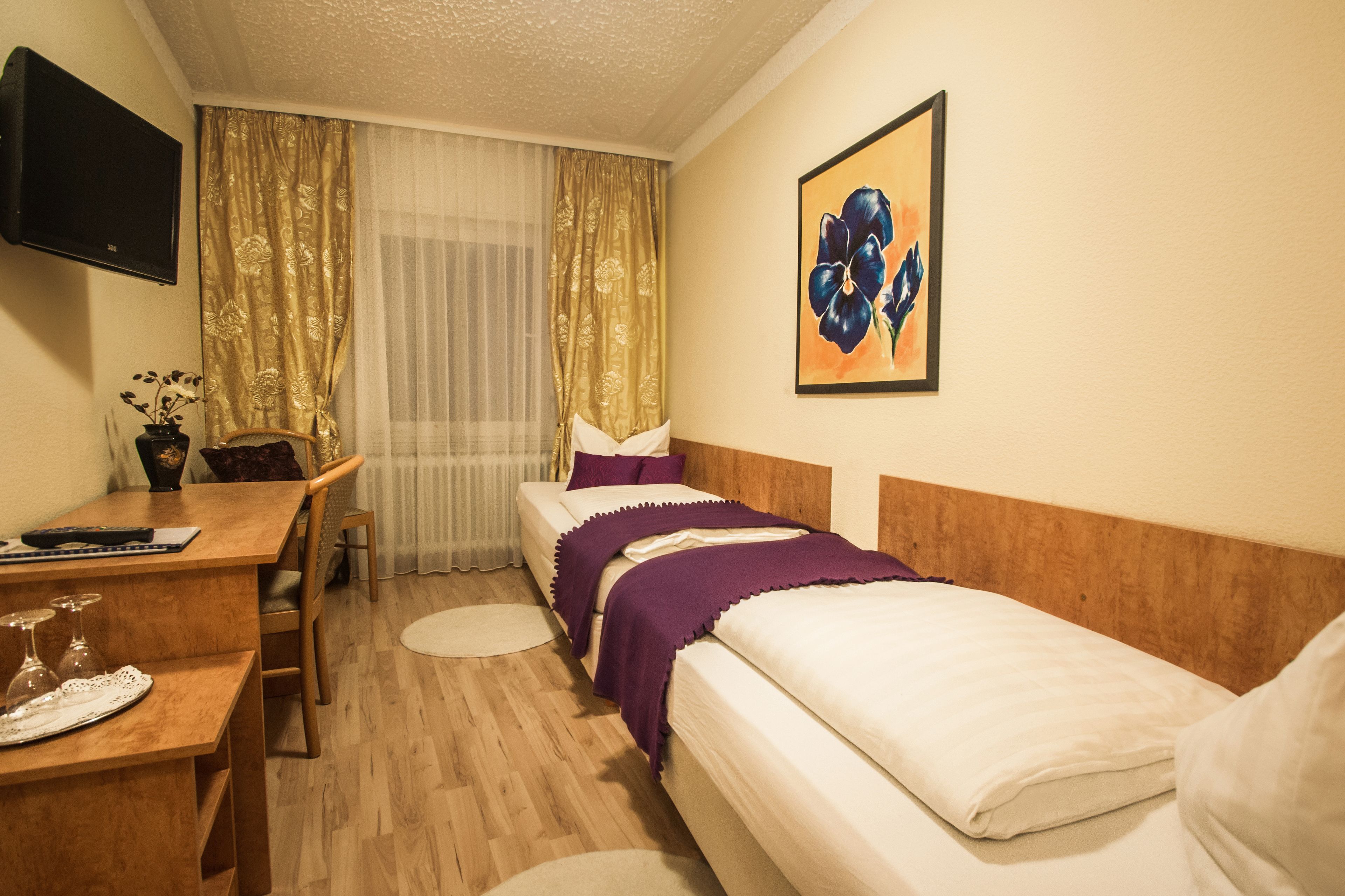 A hotel room with a bed, a desk with a chair, a TV mounted on the wall, and a window with curtains.
