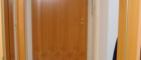 Un pasillo estrecho con una puerta de madera al final, suelo de baldosas y un abrigo colgado en la pared.