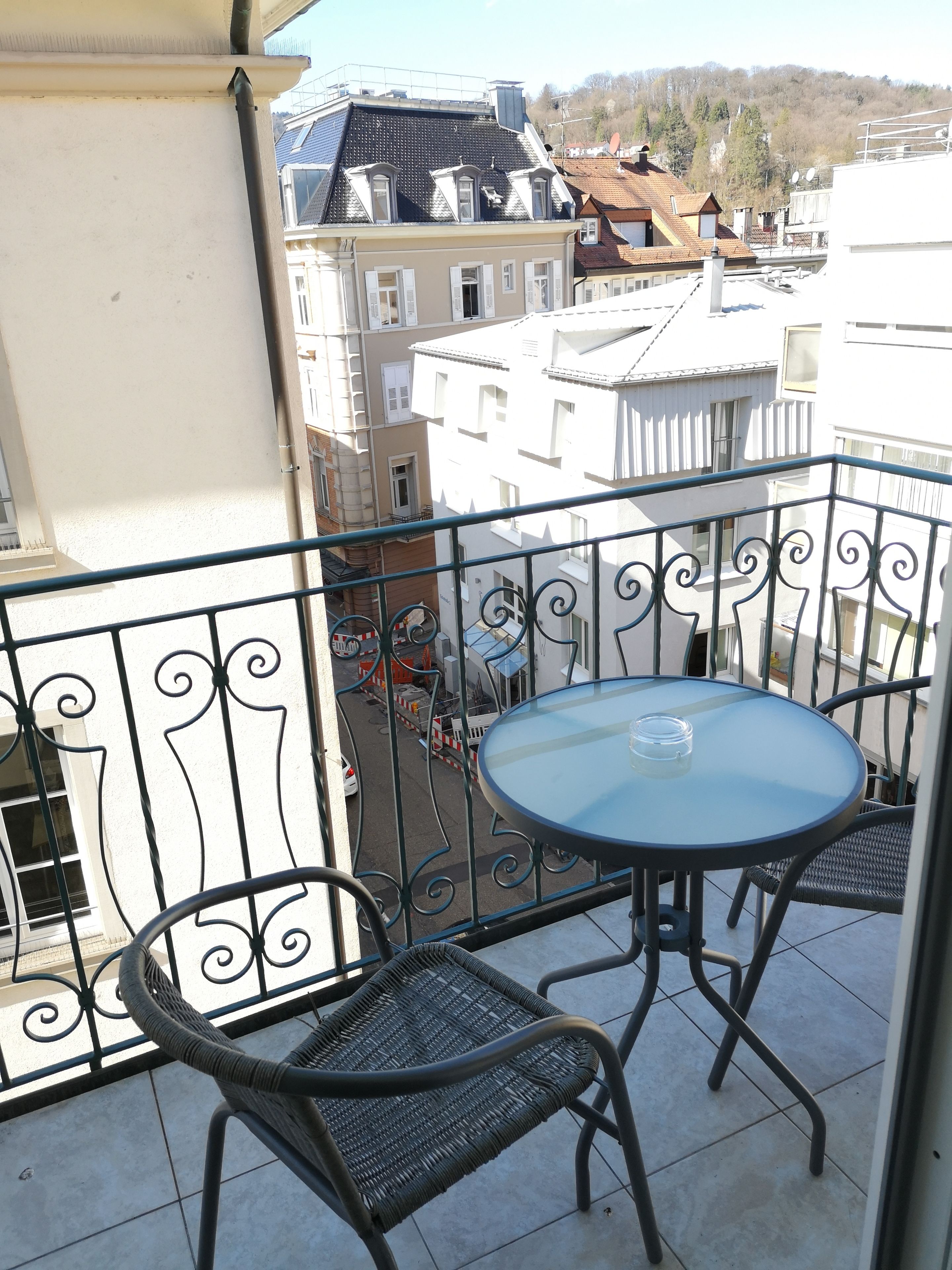 A balcony with a round table and two wicker chairs, overlooking a street with buildings and a hilly landscape in the background.