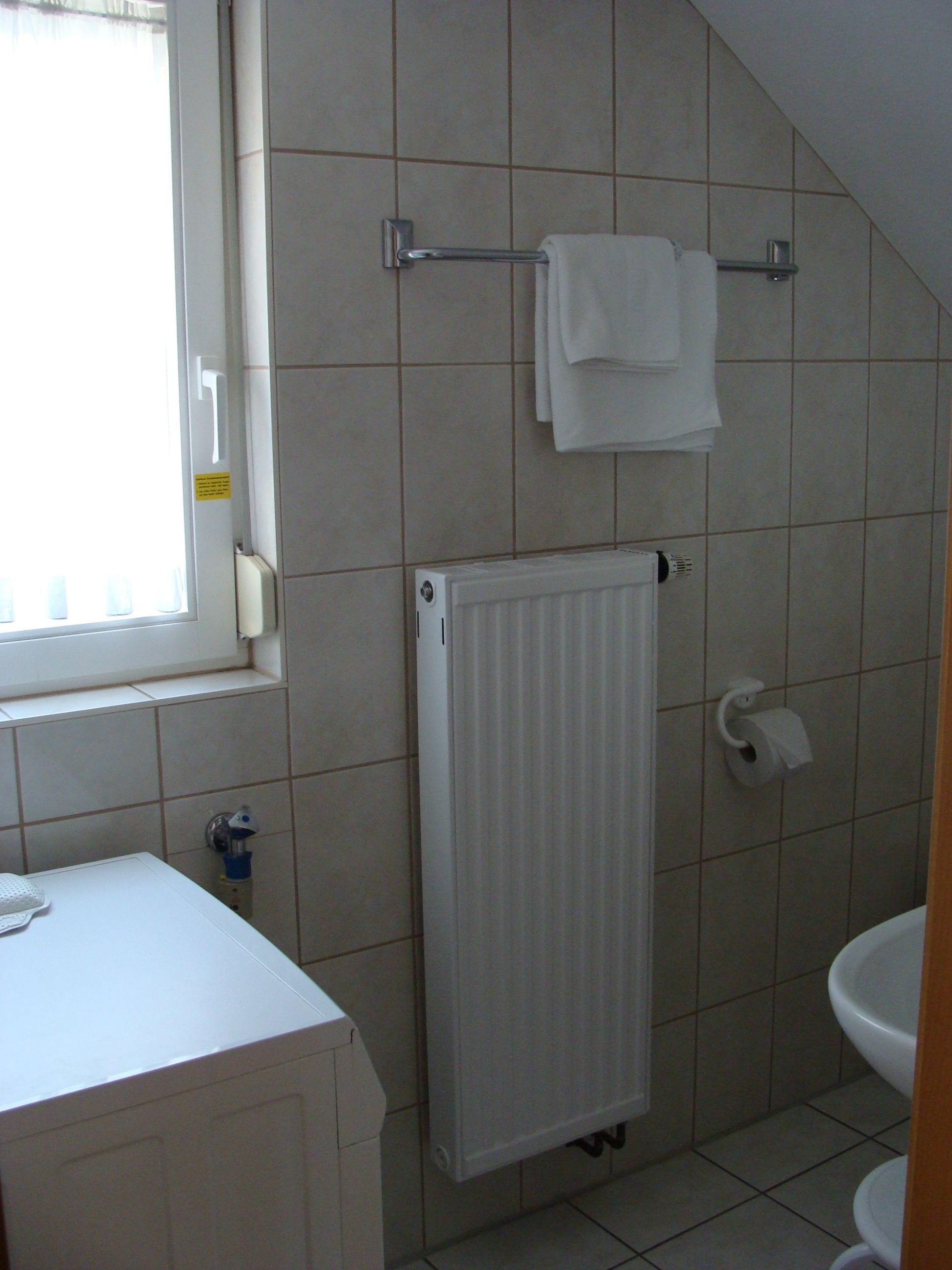 Un baño con lavamanos blanco, inodoro, toallero con toallas, radiador y ventana.