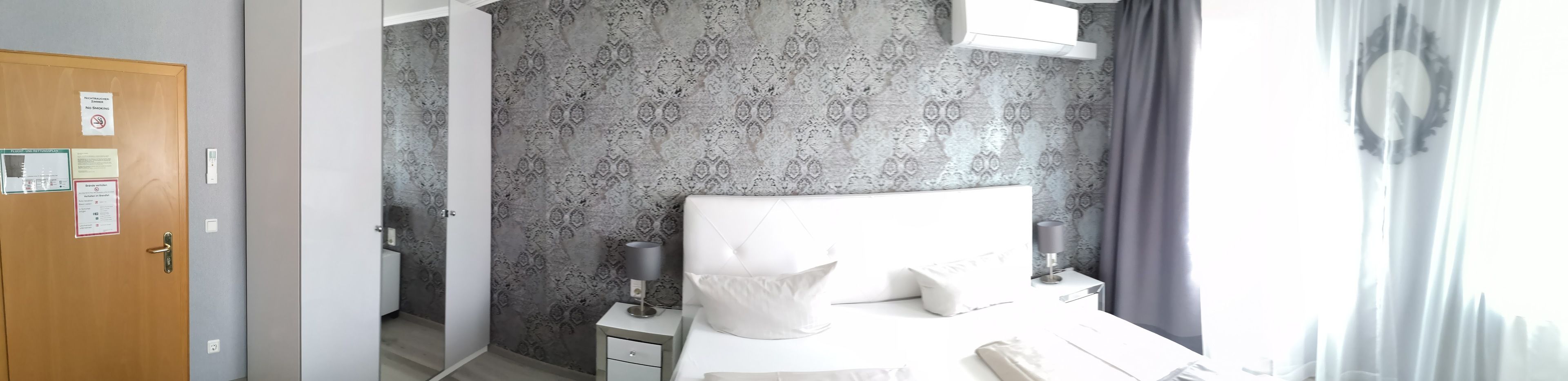 A hotel room with a patterned wallpaper, a bed with white linens, a bedside table with a lamp, and a door with a "No Smoking" sign.