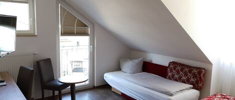 Una habitación pequeña y bien iluminada con una cama, un escritorio y una silla. Hay una ventana que ofrece luz natural y vistas al exterior.