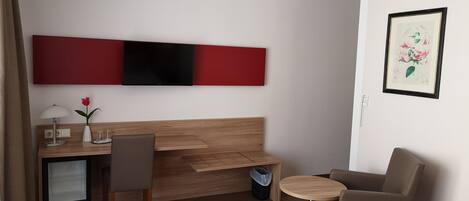 Une chambre d’hôtel moderne avec un bureau en bois, un téléviseur fixé au mur, une petite table ronde et un fauteuil confortable.