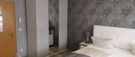 A bedroom with a bed, a wardrobe, a mirror, a bedside table with a lamp, and a wall with a patterned wallpaper.